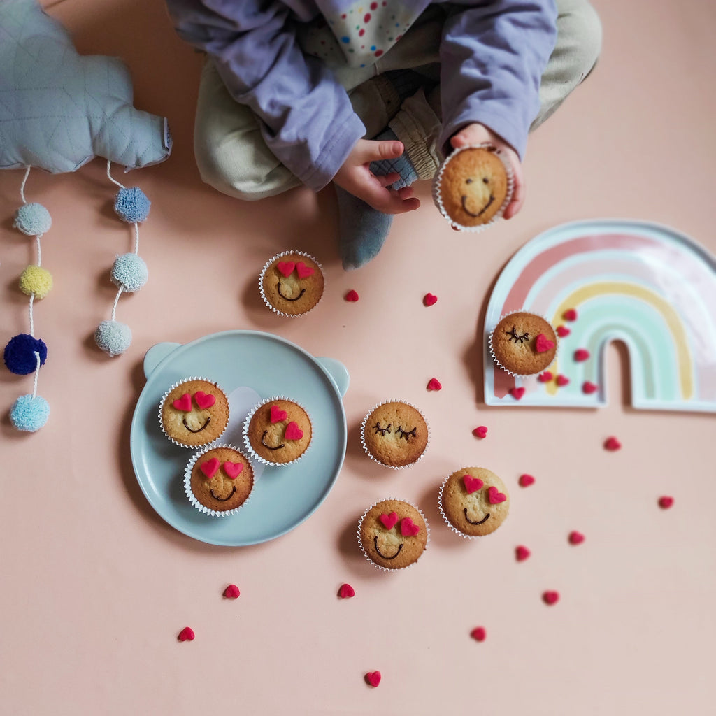 A toddler sitting on a blush pink Studio Huske silicone play mat, holding a smiley-face muffin, photographed by Game of Toddlers