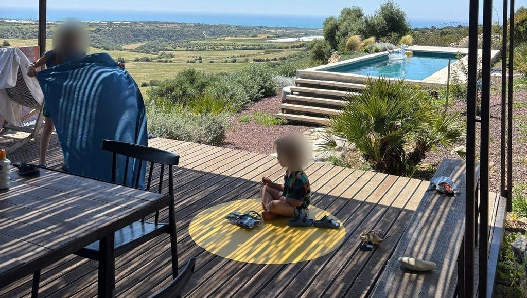 Studio Huske yellow round mat: child on a holiday terrace deck under a reed pergola with pool and sea view