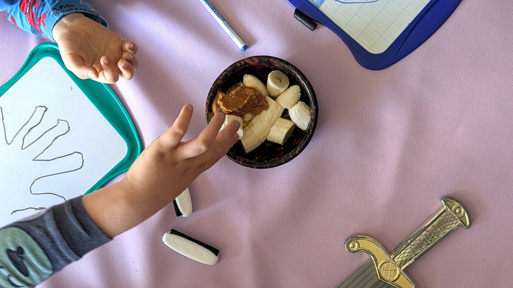 Studio Huske lilac Roam Round splat mat on the sofa: banana and peanut butter in a bowl, a hand reaching in