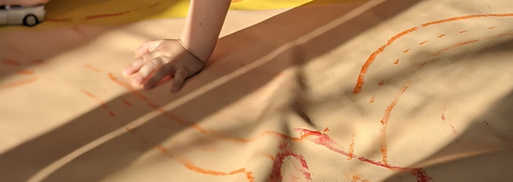 A child's hand and red and orange crayon marks on a blush Roam Square play mat on a wooden floor in evening sun