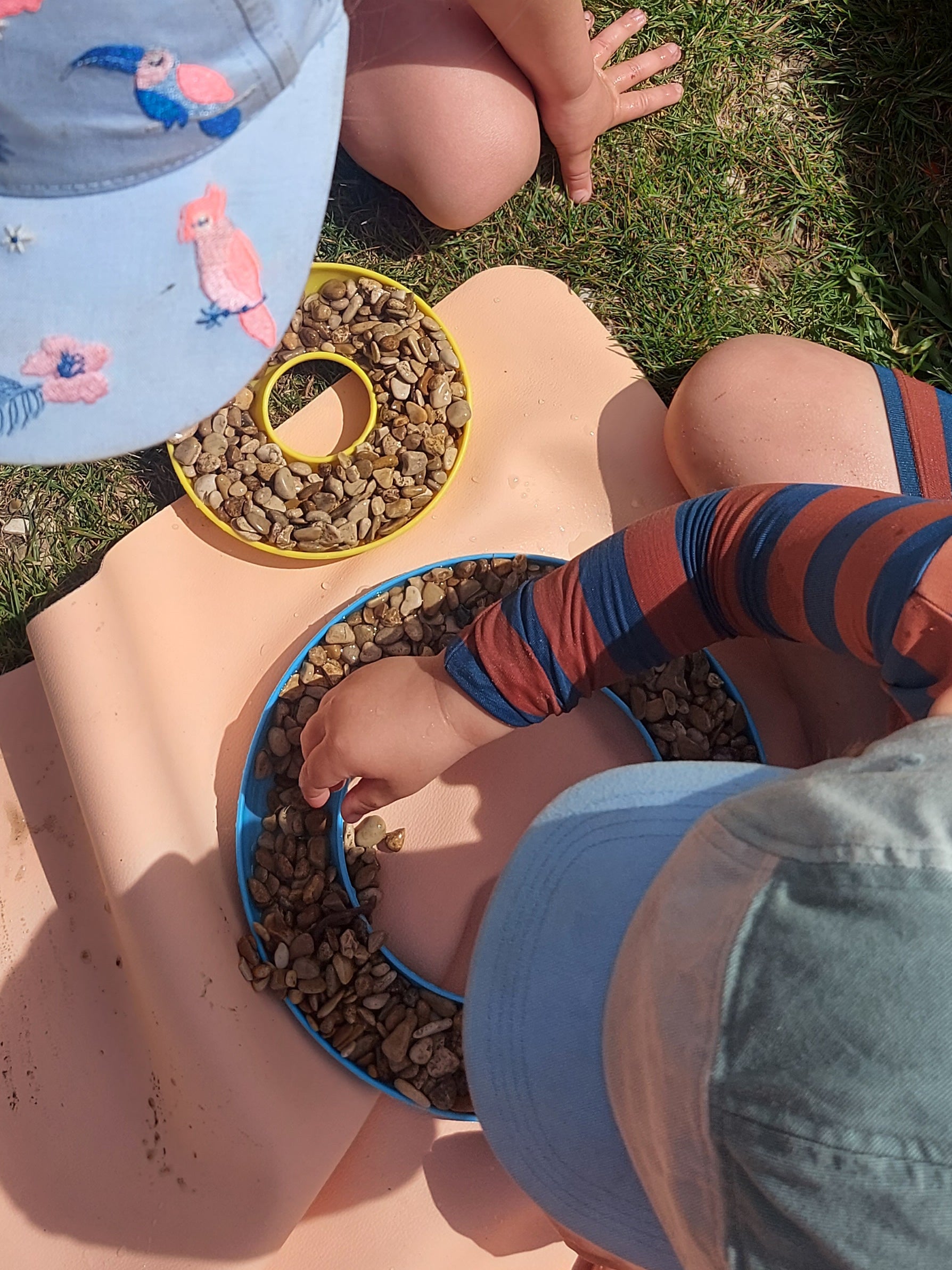 Two toddlers playing with pebbles on blush Huske mat outdoors on grass