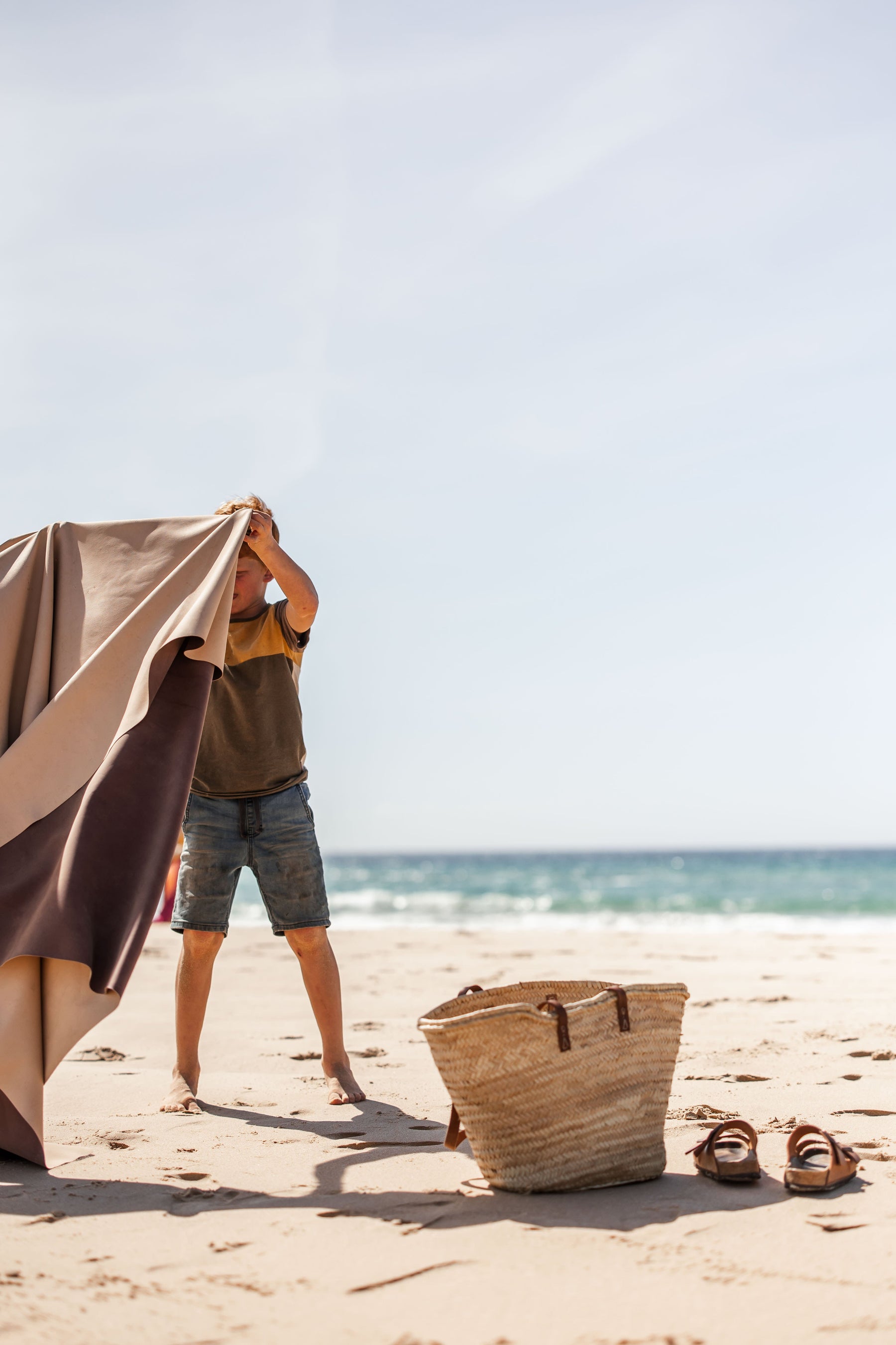 Boy unfolding Huske mat on sandy beach, straw bag and sandals beside him