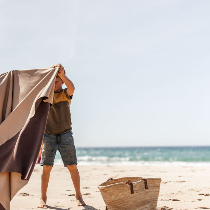 Boy unfolding Huske mat on sandy beach, straw bag and sandals beside him