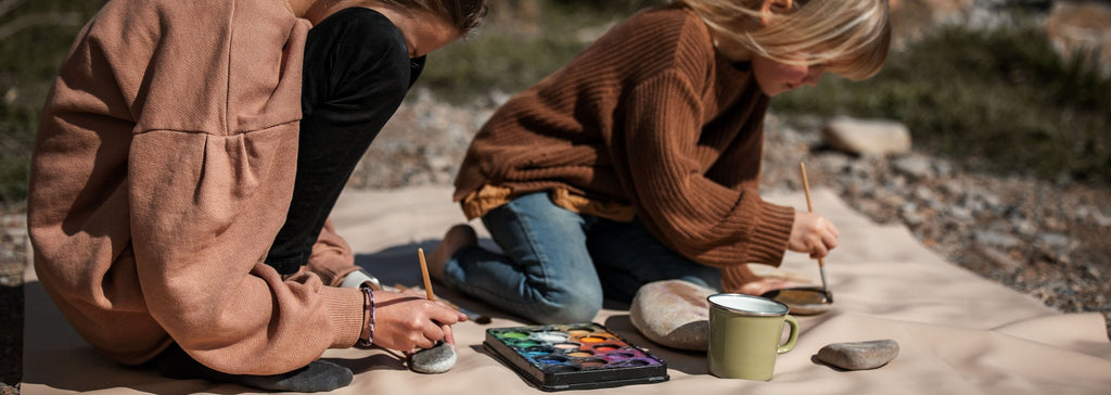 Studio Huske silicone play mat: two children painting stones on a beige mat on a pebble beach
