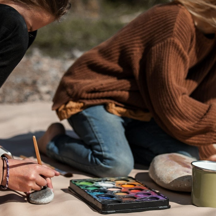 Studio Huske silicone play mat: two children painting stones on a beige mat on a pebble beach