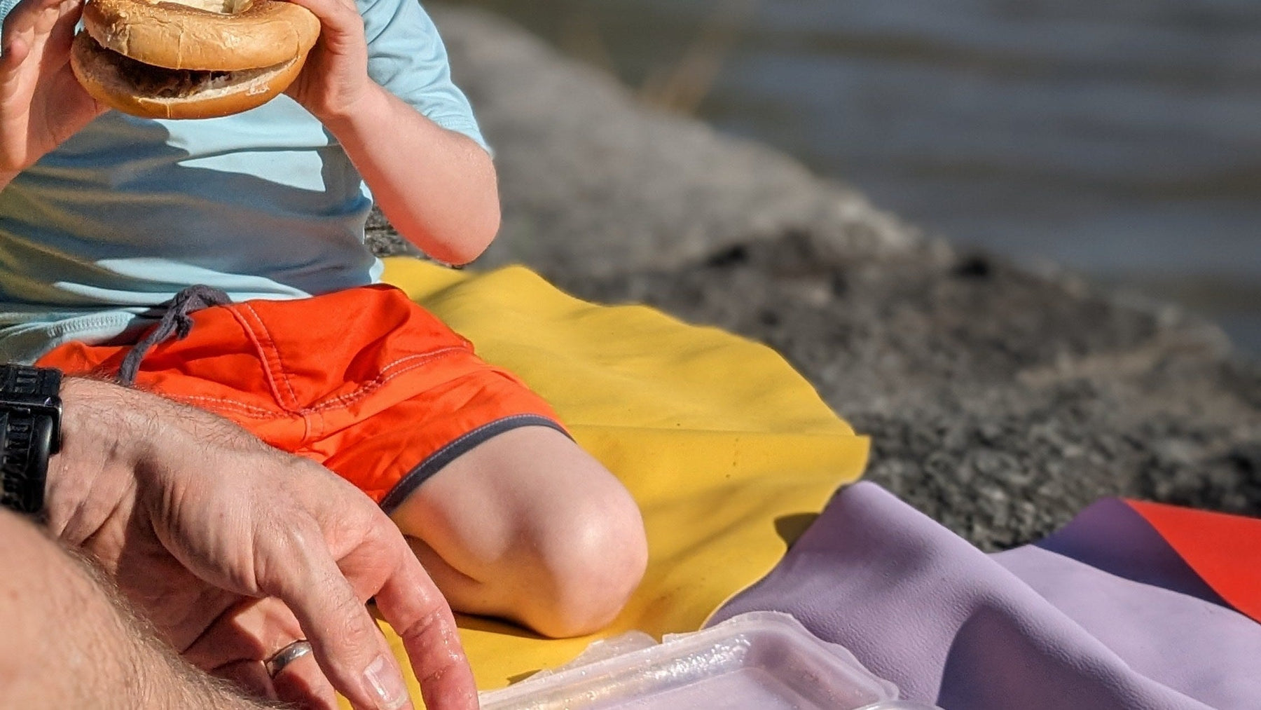 Child in orange shorts sitting on yellow and lavender Huske mats by a Swiss lake