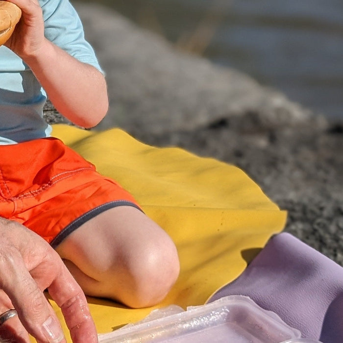 Child in orange shorts sitting on yellow and lavender Huske mats by a Swiss lake