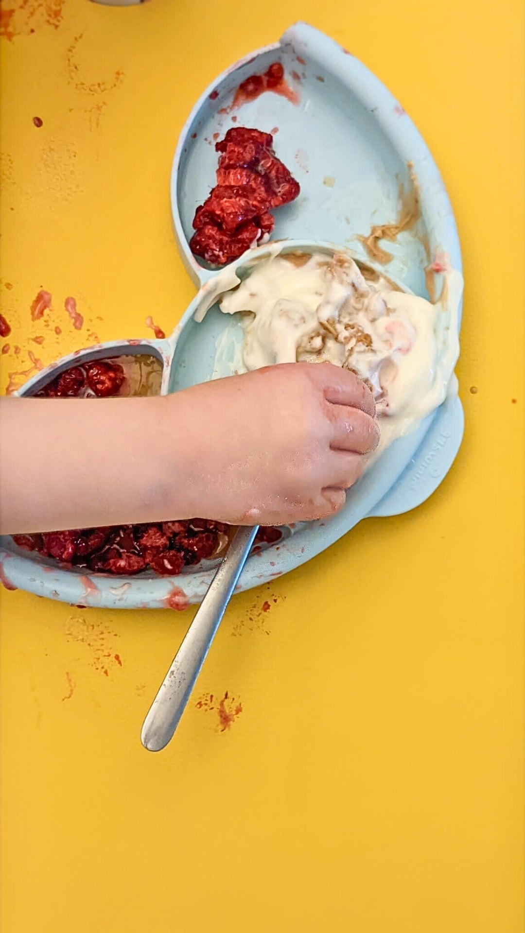 Overhead close-up of child's messy hands with raspberry yogurt on a yellow Huske silicone splat mat