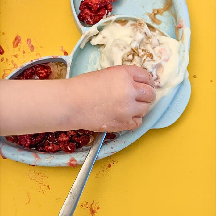 Overhead close-up of child's messy hands with raspberry yogurt on a yellow Huske silicone splat mat