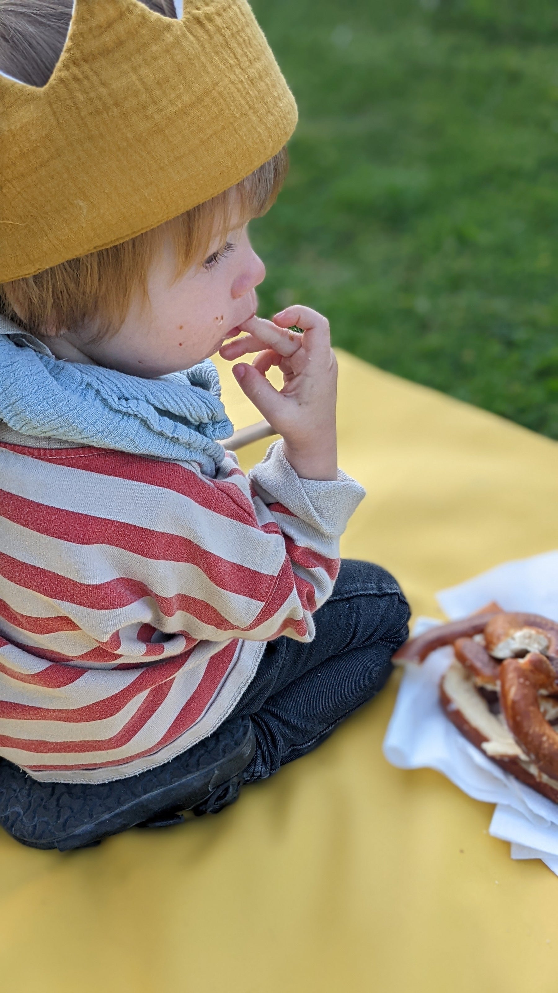 Toddler wearing crown eating Swiss pretzel on yellow Huske mat outdoors