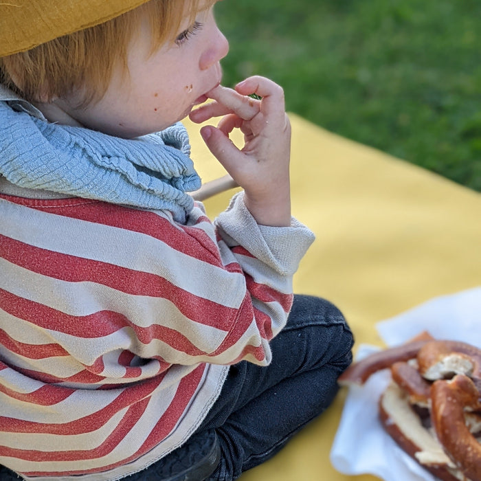 Toddler wearing crown eating Swiss pretzel on yellow Huske mat outdoors