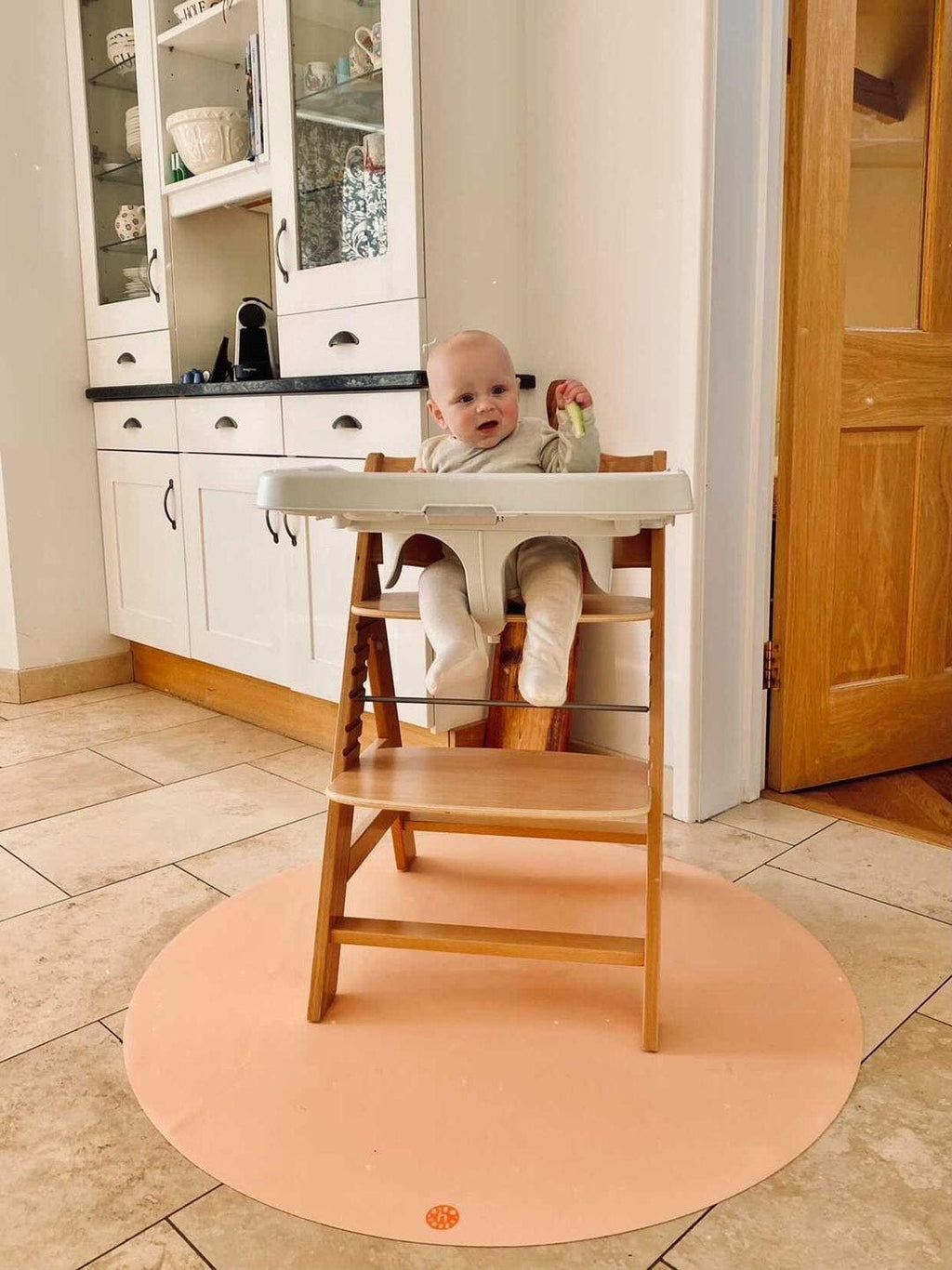 Baby smiling in wooden highchair during weaning, with   Studio Huske round silicone splat mat on kitchen floor