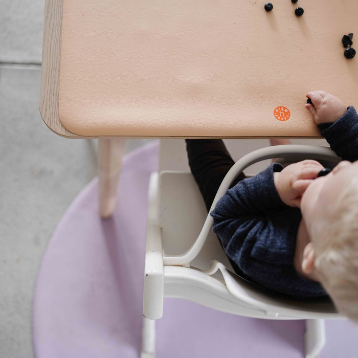 Top-down view of baby in high chair with peach Huske Wriggle mat on tray and lilac Roam round mat on floor below