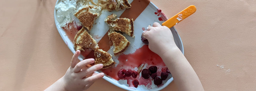 Kleinkindhände mit Pancakes und Beeren auf einem Teller, darunter eine abwischbare Matte in Blush