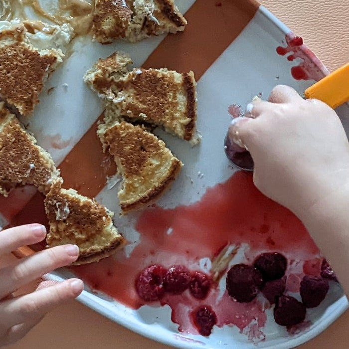 Kleinkindhände mit Pancakes und Beeren auf einem Teller, darunter eine abwischbare Matte in Blush