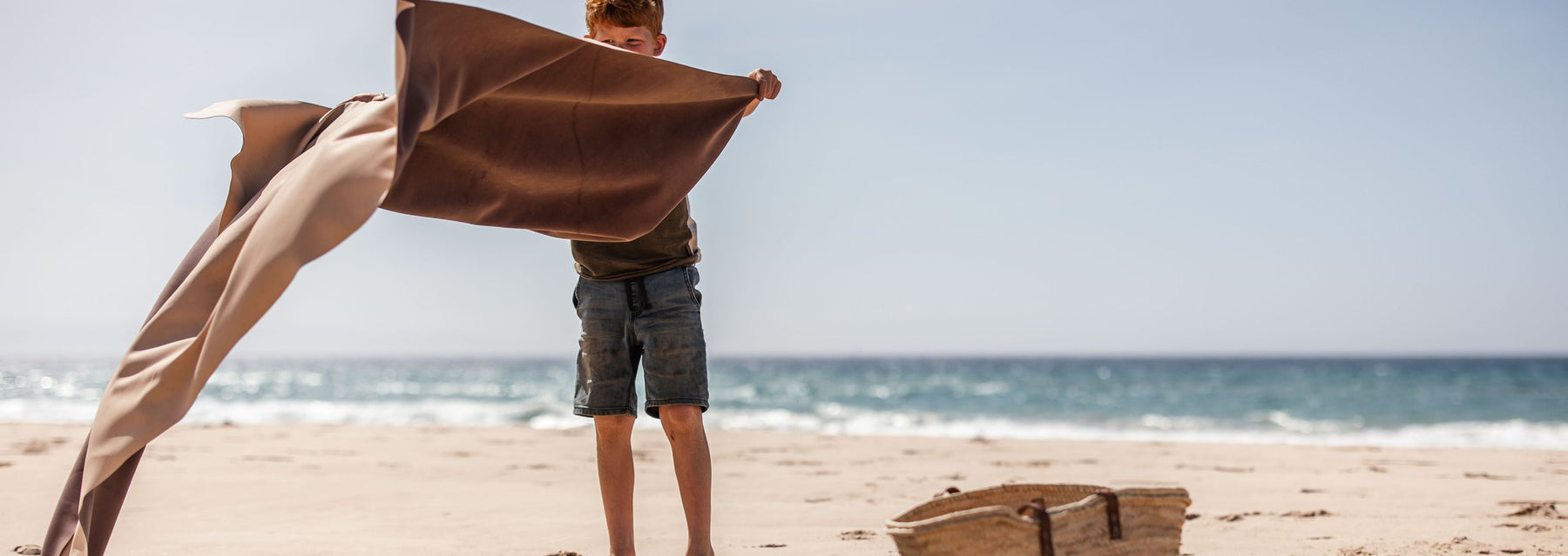 A boy shakes a mat open on a wide sandy beach, sea behind, a straw basket on the sand