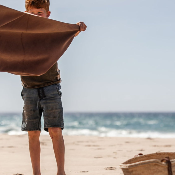 A boy shakes a mat open on a wide sandy beach, sea behind, a straw basket on the sand