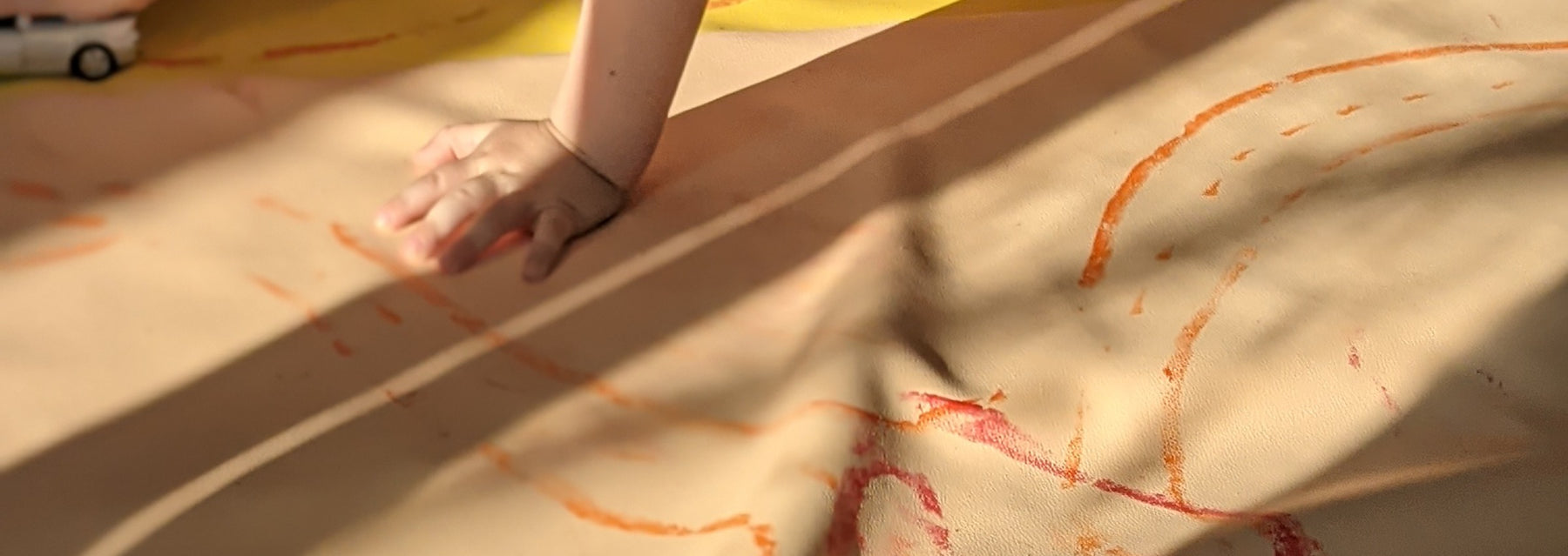 A child's hand and red and orange crayon marks on a blush Roam Square play mat on a wooden floor in evening sun