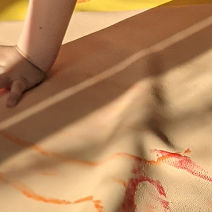 A child's hand and red and orange crayon marks on a blush Roam Square play mat on a wooden floor in evening sun
