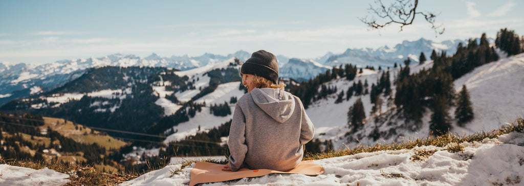 Child in a grey hoodie and beanie sitting on a peach Studio Huske mat on a snowy alpine ridge, looking out over the Swiss Alps