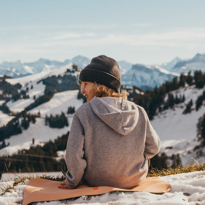 Child in a grey hoodie and beanie sitting on a peach Studio Huske mat on a snowy alpine ridge, looking out over the Swiss Alps