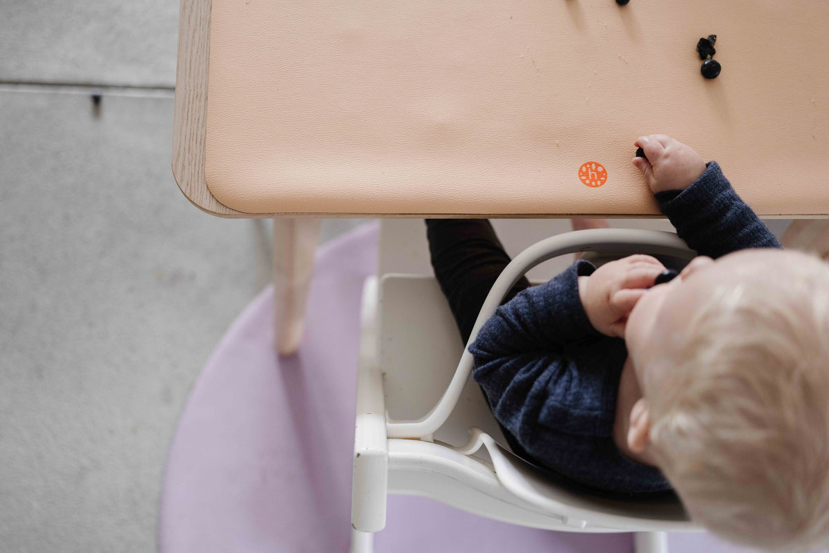 Toddler in high chair eating blueberries with peach silicone mat on tray and lilac splat mat on floor below
