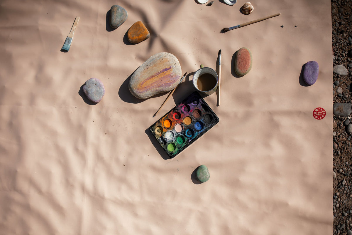 Overhead of painted pebbles and art supplies spread on beige silicone craft mat
