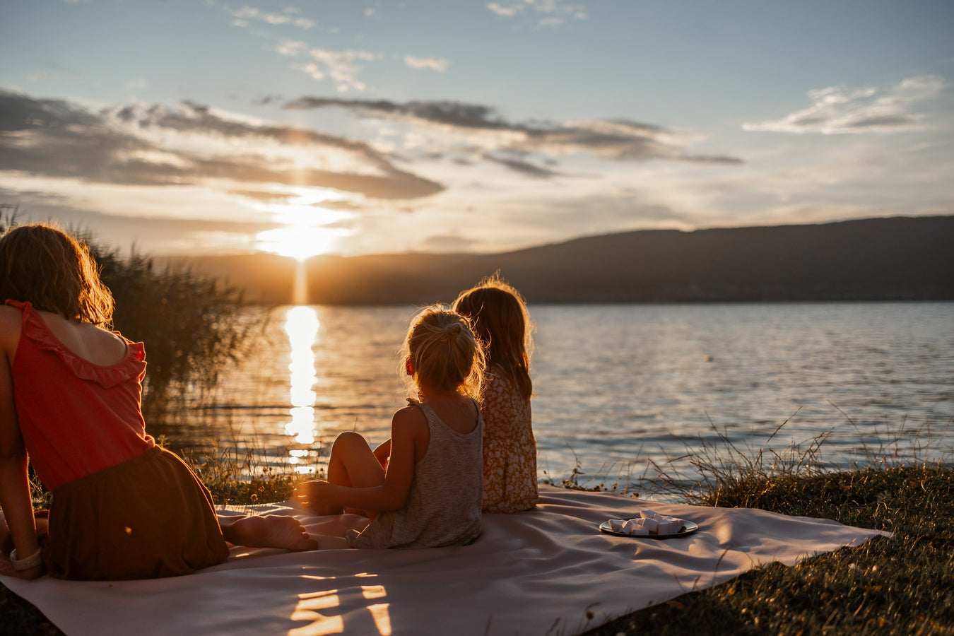 Family on beige silicone mat watching sunset by Swiss lake with mountain panorama