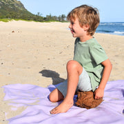Gallivant XL Lilac/Red silicone family picnic mat, side profile of smiling boy on lavender huske mat holding coconut