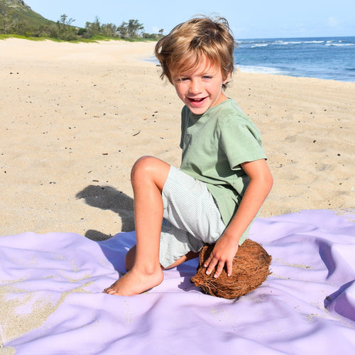 Gallivant XL Lilac/Red silicone family picnic mat, smiling boy sitting on lavender huske mat at the beach holding a whole