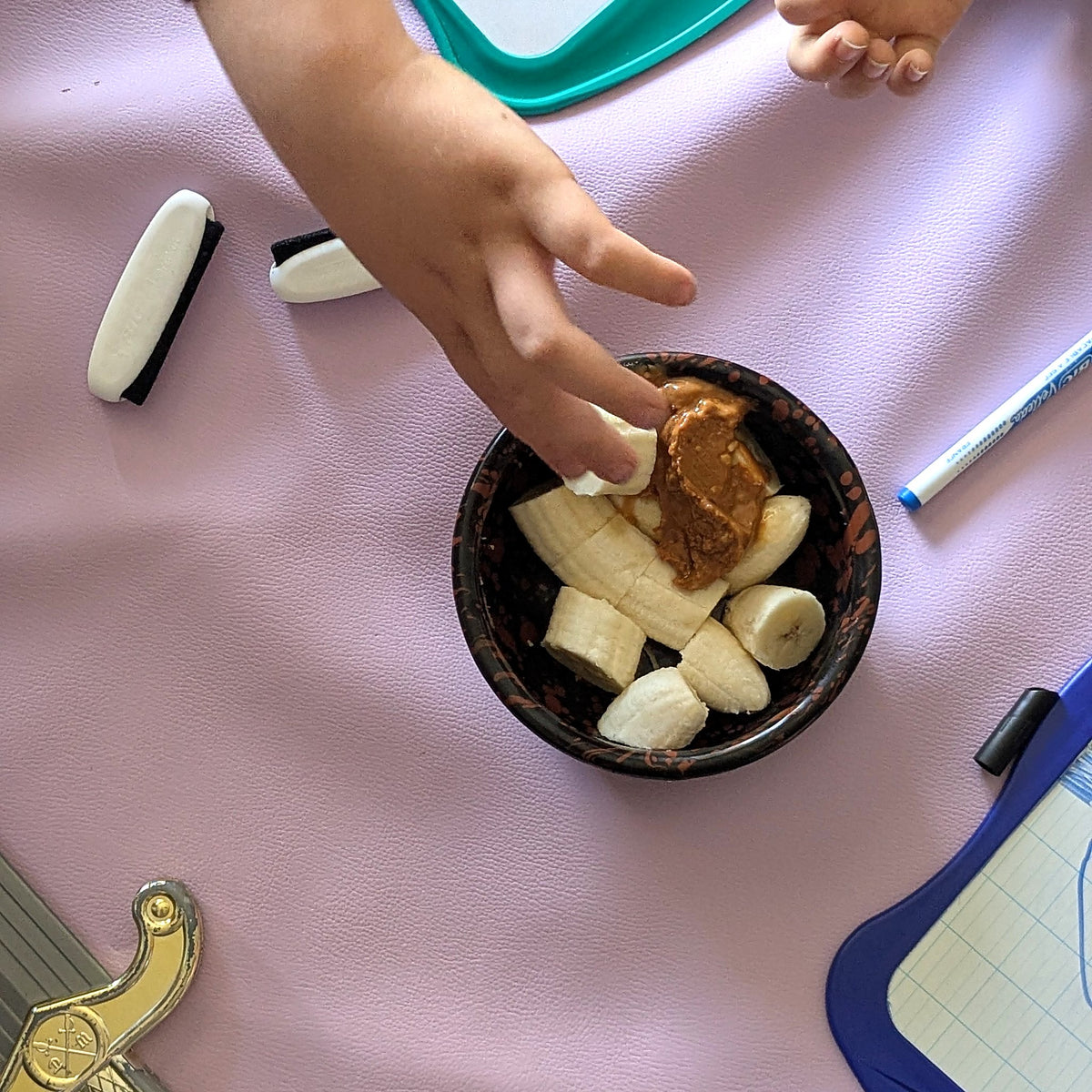 Roam Round Lilac/Red silicone splat mat on the sofa, a hand taking banana from the snack bowl beside drawing boards