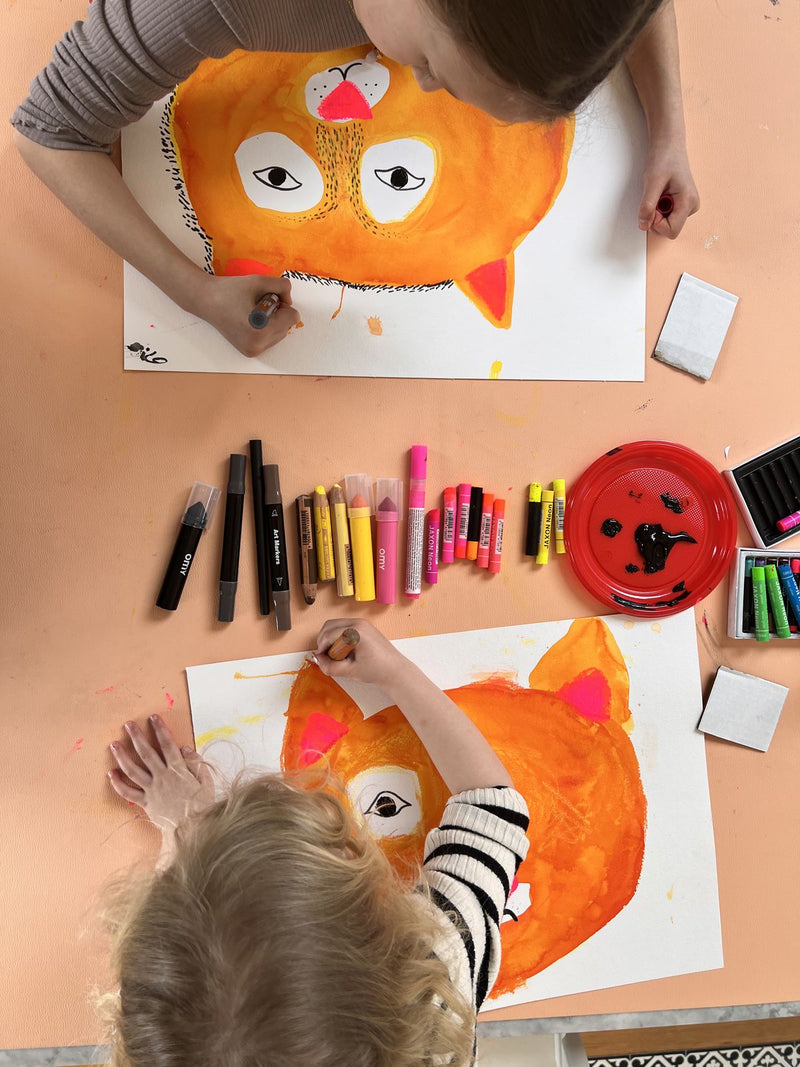 Children painting large orange tiger faces on paper on a peach Huske mat covered with art materials