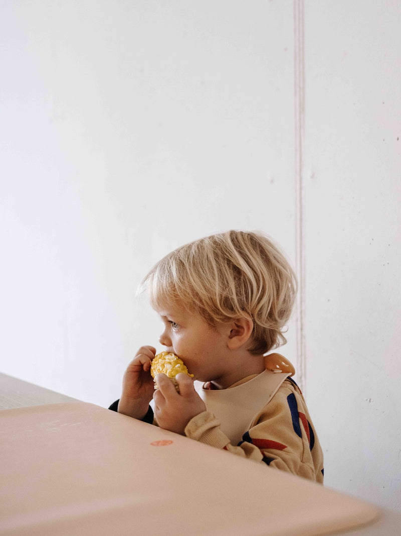 Studio Huske silicone placemat: toddler eating corn at a table on a peach Wriggle mat with the huske logo