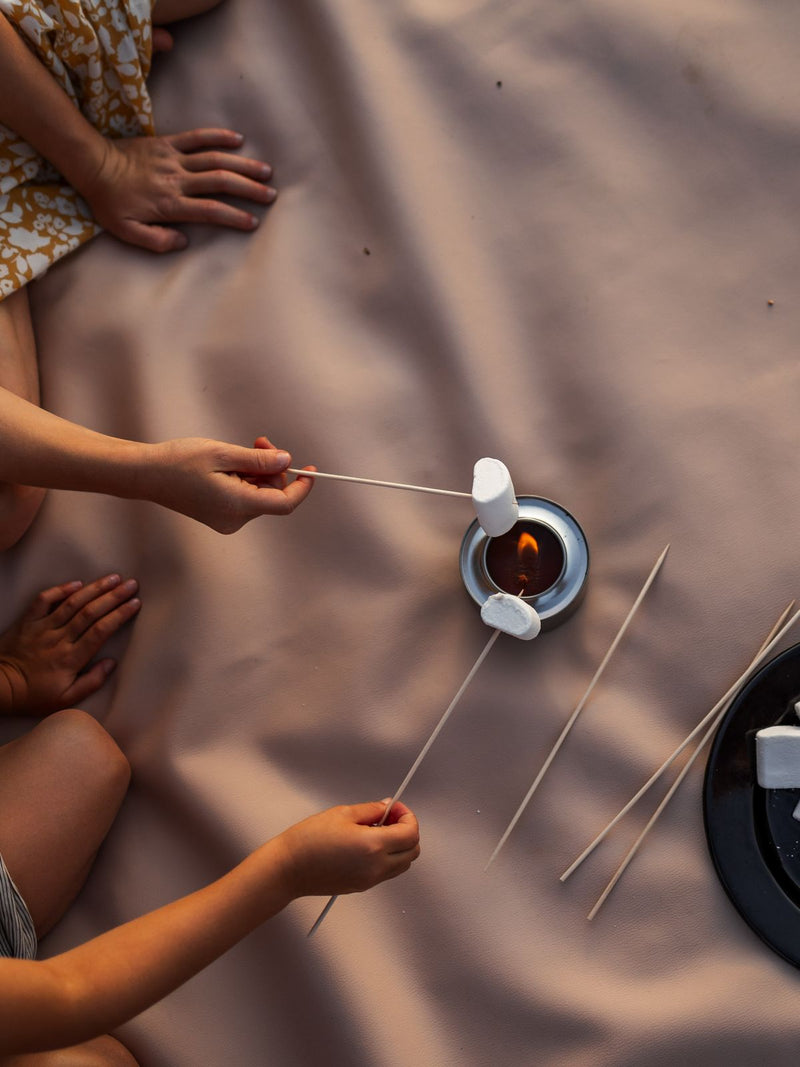 Children toasting marshmallows over a small flame on a beige Huske mat by the lake