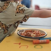 Child reaching for a plate of pasta on a waterproof silicone leather changing mat, Wriggle Mat (S).